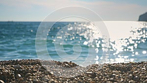 Closeup detail - small pebbles or rough sand on a beach, blurred afternoon sun lit calm sea in background, camera slide to side