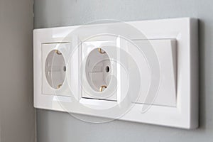 A closeup diagonal view of a group of white european electrical outlets and a switch located on a painted gray wall