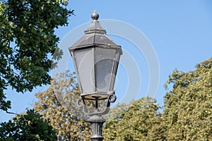 Closeup of a dirty street lantern captured at daylight against a blue sky and leafy trees
