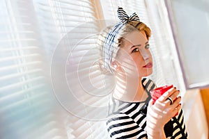 Closeup on elegant beautiful blond pinup young woman drinking coffee standing at white blinds sun lighted window background