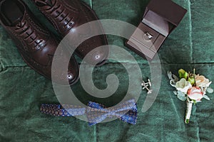 Closeup of elegant stylish dark male accessories on green background. Top view of bow-tie, shoes, floral corsage, golden