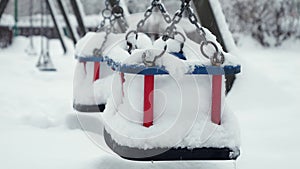 Closeup of empty chain swings on public playground at park being covered with snow during snowfall. Concept of winter holidays.