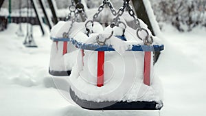 Closeup of empty chain swings on public playground at park being covered with snow during snowfall. Concept of winter holidays,