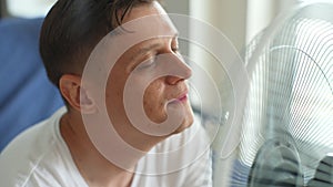 Closeup face of happy young man enjoying cool wind from electric fan at home at summer heat. Male suffer from heat high