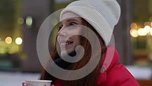 Closeup face of happy young woman in winter clothes holding and drinking hot coffee from takeaway cup outdoors in cold