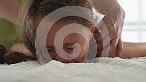 Closeup face suffering from pain of adorable little girl having back shoulder massage by unrecognizable female pediatric