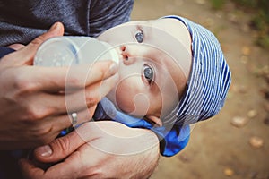 Closeup of father`s hands gives his infant son milk from a bottle outdoors. Baby drinks milk. Childcare concept.