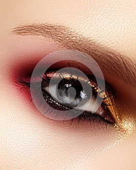 Closeup female eye with fashion bright make-up. Beautiful gold, red eyeshadow, glitter, black eyeliner. Shape Eyebrows