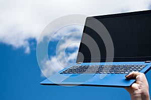 Closeup of female hand holding a blue laptop against the blue sky with white clouds