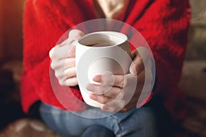 Closeup of female hands with a cup of beverage. Beautiful girl in red sweater holding cup of tea in the morning sunlight