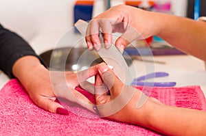 Closeup females hands getting manicure treatment from woman in salon environment, pink towel surface, blurry background