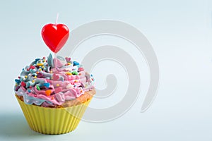 Closeup festive creamy cupcake with sprinkles and candle in form of red heart on light background