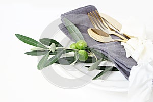 Closeup of festive table summer setting with golden cutlery, olive branch, grey linen napkin, porcelain dinner plate and photo