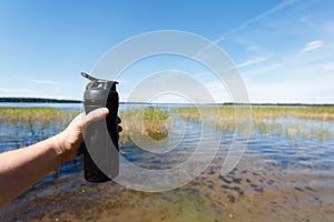 Closeup of fitness shaker bottle in the men's hand against the beautiful lake. Sports snack the fresh air. The concept of proper