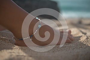 Closeup of girl right foot with boho style bracelet on sand, model sits on beach