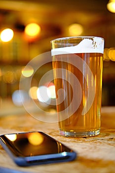 Closeup of a glass of beer on the table near a phone under the lights with a blurred background