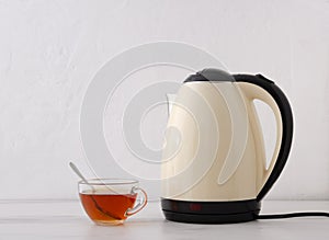 Closeup of glass cup of black tea and bright electric kettle on the table against white wall