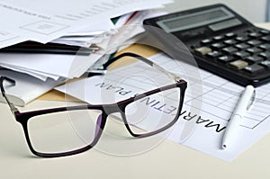 Closeup of glasses, blank paper for marketing plan, pile of documents, calculator on the desk