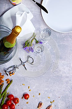 Closeup of a gray table with plate, a bottle of champagne, tomatoes, asparagus, glasses, corkscrew on a gray background.