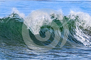Closeup of green ocean wave curling. White foam and spray on top; blue ocean behind.