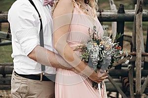 Closeup of groom embracing a bride in beautiful pink dress. Woman holding floral bouquet in hands. Boho wedding and