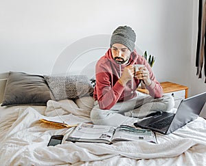 Closeup of a guy sitting on the bed with his laptop and books and a cup of coffee in his hands