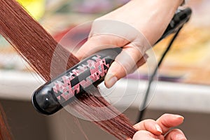 Closeup of hairdresser hands with styling iron straightening woman hair in salon.