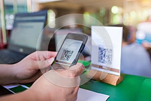 Closeup of a hand holding phone and scanning QR code with blurry cashier at counter service. Man hands paying with QR code.