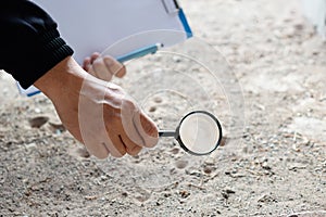 Closeup hand holds magnifying glass to explore tiny insects in holes on ground.