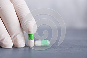 Closeup hand in medical latex glove holding two green and white capsules on gray table