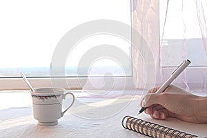 Closeup of hand and pen, writing on notebook before window, coffee cup on table, copy space