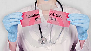 Closeup, hands in blue medical gloves hold and tear red paper card with an inscription Coronary artery disease. doctor