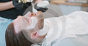 Closeup hands of female barber, who applies with brush shaving cream on young bearded man's face and neck