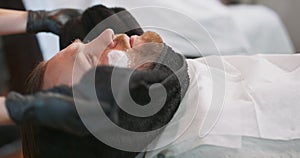 Closeup hands of female barber, who wraps a black towel on young bearded man's face and neck, with shaving cream on