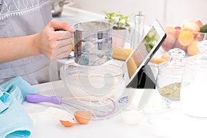 Closeup hands lady standing in kitchen and cooking the dough. Looking at tablet computer. Female following recipe on