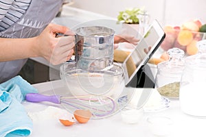 Closeup hands lady standing in kitchen and cooking the dough. Looking at tablet computer. Female following recipe on