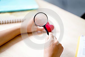 Closeup hands of medical student holding a magnifying glass on a yarn red heart and blurred background.