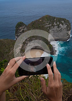 Closeup hands with mobile phone doing photo of Kelingking Beach cliffs in Nusa Penida, Bali