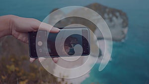 Closeup hands with mobile phone doing photo of Kelingking Beach cliffs in Nusa Penida, Bali