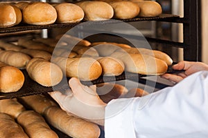 Closeup of hands of professional chef man in white uniform standing near shelves full with fresh bread and pulling baking tray for