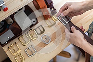 Closeup hands of professional tanner choosing belt buckle on wooden table at leather workshop