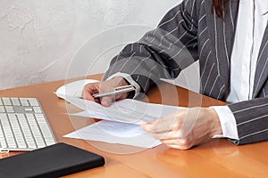 Closeup hands of woman in formal suit and white shirt, holding pen, sheets of paper, near computer keyboard, mouse and notebook.