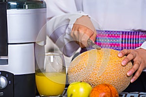 Closeup hands of woman holding small knife cutting into honey melon, glass on the table next to it, healthy lifestyle