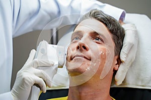 Closeup handsome man having color light therapy to stimulate facial skin by professional cosmetologist wellbeing