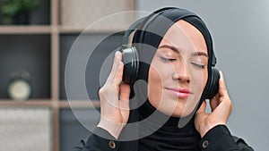 Closeup happy face of Muslim woman in black headscarf listening music in headphones relaxing at home