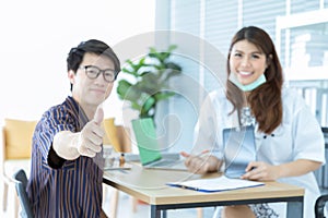 Closeup of happy young Asian male patieent with casual clothes is showing thumbs up while sitting with beautiful female doctor