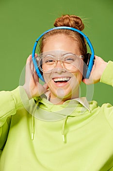 Closeup happy young girl wearing glasses and wireless headphones listening to music and smiling over green background