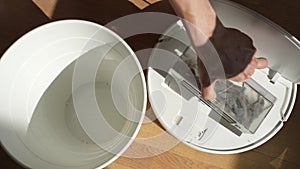 Closeup high-angle view of unrecognizable man shaking container of robot vacuum cleaner with dust and dirt in trash can