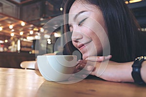 Closeup image of Asian woman sit with chin resting on her hands and closing her eyes smelling hot coffee on wooden table with feel