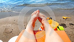 Closeup image of bare feet lying on the sun bed at sea beach against toys for playing in sand